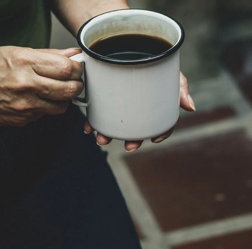 Two hands holding a white enamel mug of black coffee