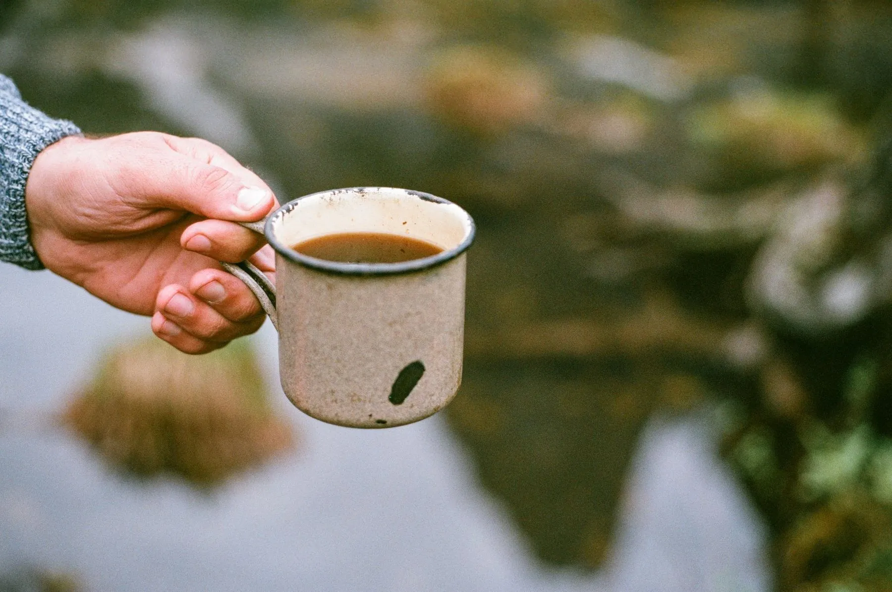 A hand holding a speckled enamel mug of coffee over a blurred lake and trees