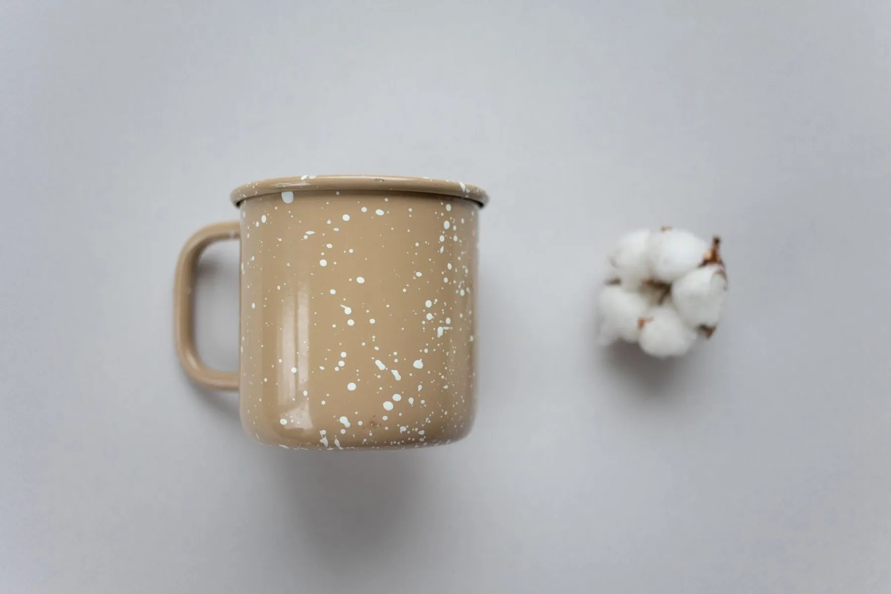 Speckled enamel mug in a warm tan glaze flecked with white, beside a cotton flower
