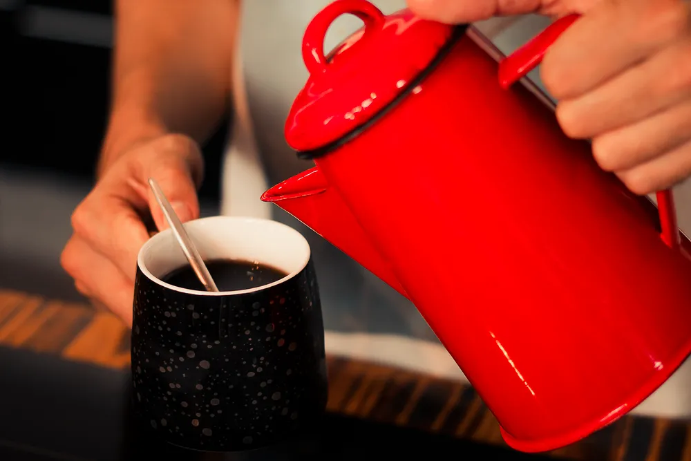 Coffee poured from a vibrant red enamel pot into a speckled cup